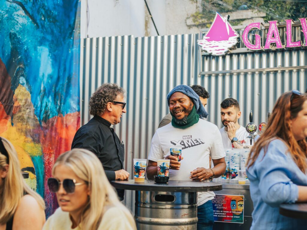 People enjoying drinks and chatting outdoors at a Galway Hooker Secret Sessions event, with colourful graffiti artwork and a pink Galway Hooker sign in the background. A group gathers around a table made from a beer keg, creating a relaxed, social atmosphere.
